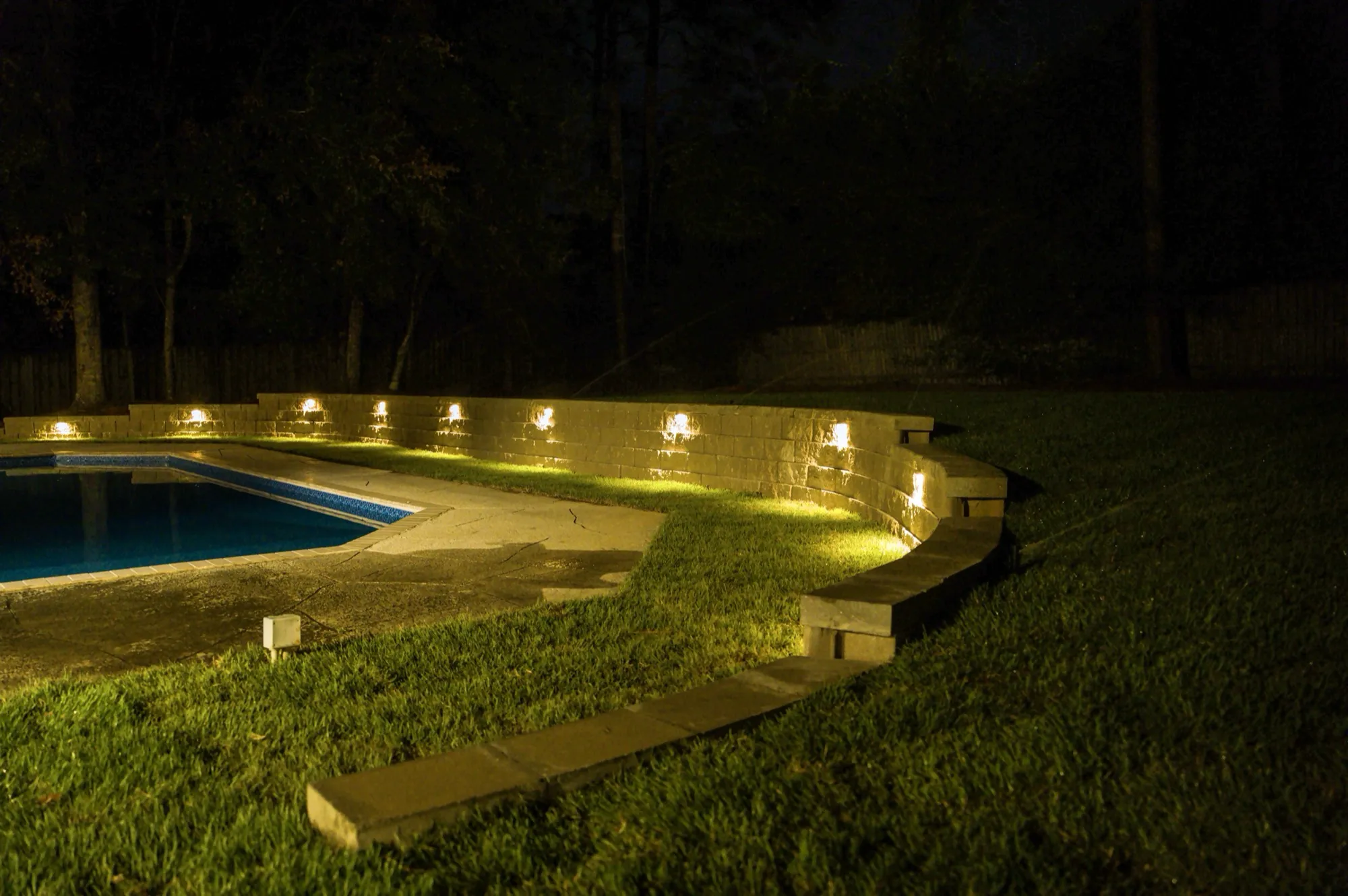 Curved retaining wall with low-voltage uplighting glowing along a backyard pool at night in Baldwin County, Alabama